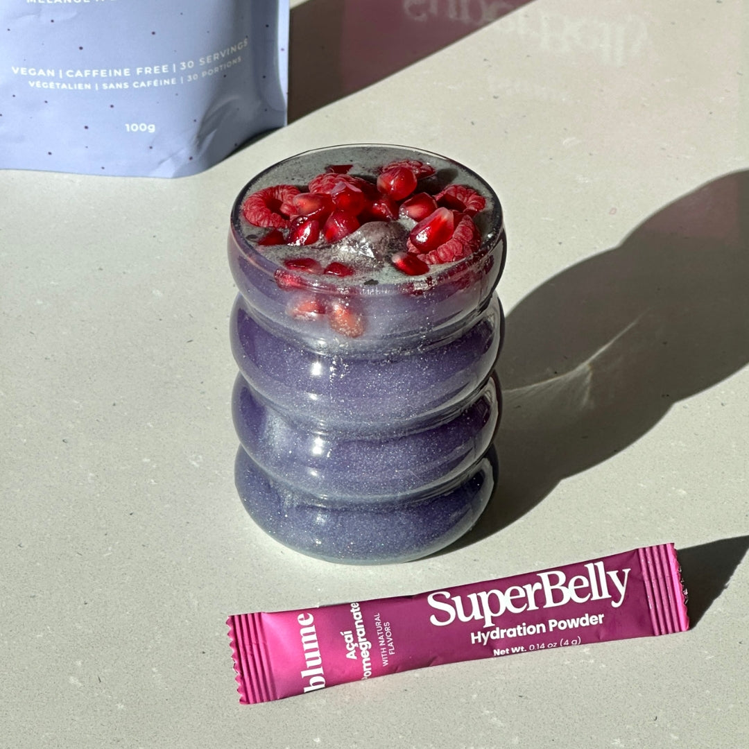 A wavy glass holds a purple smoothie topped with pomegranate seeds. Beside it, there is a pink sachet of SuperBelly Hydration Powder with acai and pomegranate flavor. In the background, there is a partially visible container.